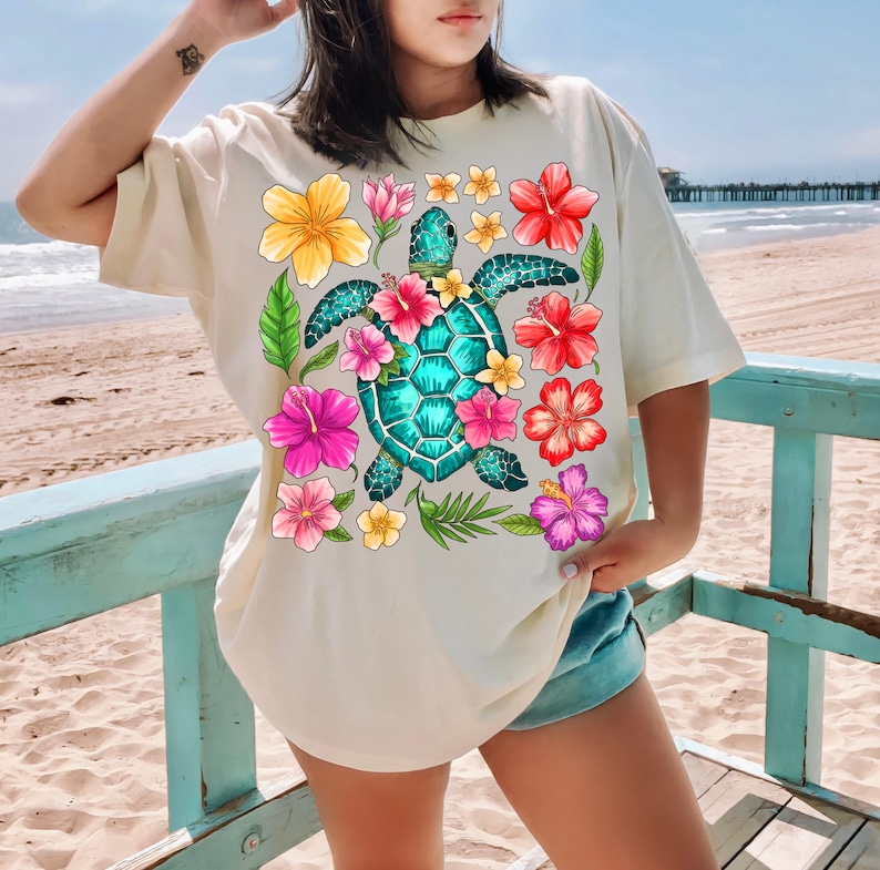 May include: A cream coloured t-shirt with a graphic of a green sea turtle surrounded by pink, red, yellow, and orange hibiscus flowers.