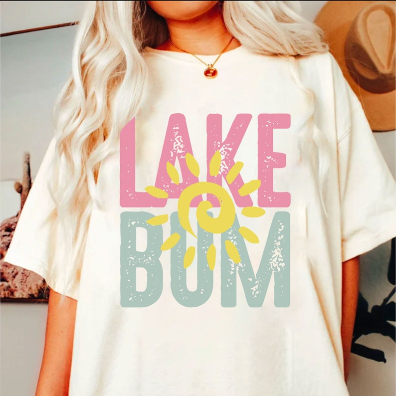 Op de afbeelding: Een wit T-shirt met de tekst "LAKE" in roze, een gele zonafbeelding en de tekst "BUM" in blauw.