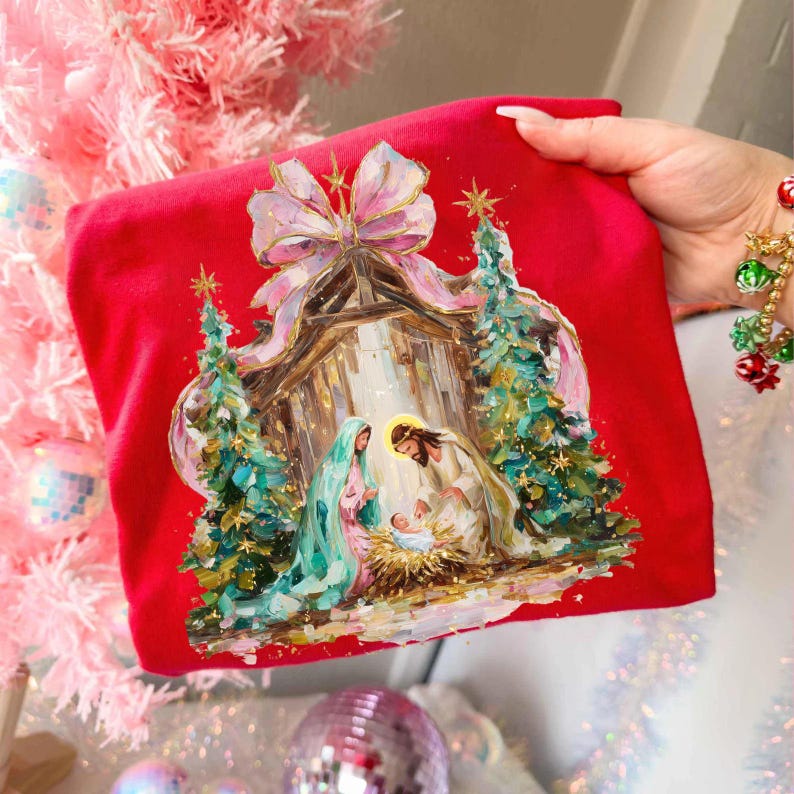 May include: Red Christmas-themed sweatshirt featuring a Nativity scene with Mary, Joseph, and baby Jesus. The design includes a wooden stable, Christmas trees, and a large bow. The artwork is detailed with gold accents and a soft, painterly style.