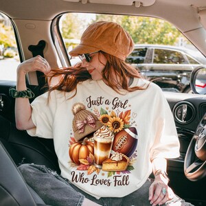 K&ouml;nnte beinhalten: Cremefarbenes T-Shirt mit dem Text "Just a Girl Who Loves Fall" und einer herbstlichen Grafik. Die Grafik zeigt einen K&uuml;rbis, einen Football, einen Latte, einen Kuchen und eine Strickm&uuml;tze.