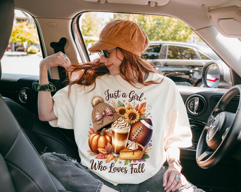 K&ouml;nnte beinhalten: Cremefarbenes T-Shirt mit dem Text "Just a Girl Who Loves Fall" und einer herbstlichen Grafik. Die Grafik zeigt einen K&uuml;rbis, einen Football, einen Latte, einen Kuchen und eine Strickm&uuml;tze.