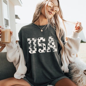 K&ouml;nnte beinhalten: Ein dunkelgraues T-Shirt mit einem wei&szlig;en "USA"-Motiv aus Sternen. Das Shirt ist &uuml;berdimensioniert und hat eine lockere Passform.