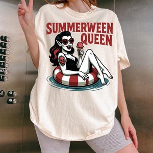 May include: Cream-colored t-shirt with the text "Summerween Queen" in red. The graphic features a vampire woman in a black swimsuit, heart-shaped sunglasses, and a skull tattoo, floating in a red and white striped inner tube, holding an ice cream cone.