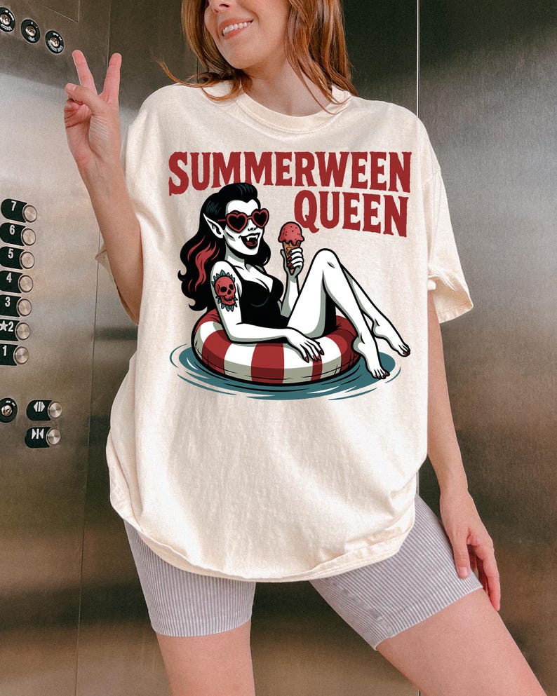May include: Cream-colored t-shirt with the text "Summerween Queen" in red. The graphic features a vampire woman in a black swimsuit, heart-shaped sunglasses, and a skull tattoo, floating in a red and white striped inner tube, holding an ice cream cone.