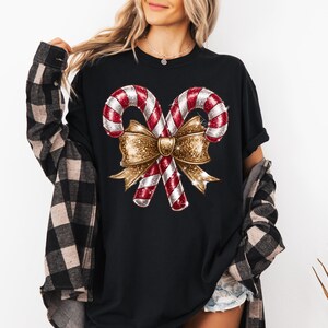 Puede incluir: Camiseta negra con un dise&ntilde;o navide&ntilde;o. El gr&aacute;fico incluye dos bastones de caramelo rojos y blancos con un lazo dorado brillante. La camiseta es de cuello redondo y tiene mangas cortas. La persona lleva una camisa a cuadros negra y marr&oacute;n.