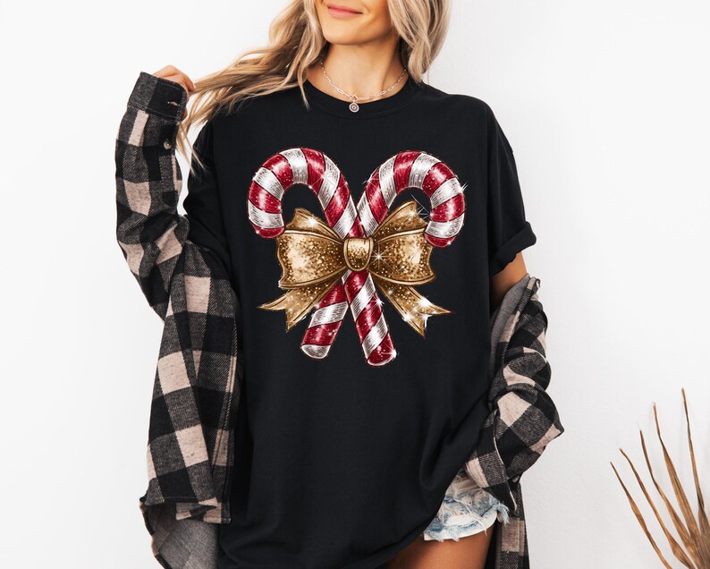 Puede incluir: Camiseta negra con un dise&ntilde;o navide&ntilde;o. El gr&aacute;fico incluye dos bastones de caramelo rojos y blancos con un lazo dorado brillante. La camiseta es de cuello redondo y tiene mangas cortas. La persona lleva una camisa a cuadros negra y marr&oacute;n.