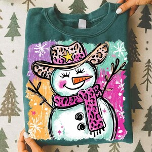 May include: A teal sweatshirt with a playful snowman illustration. The snowman is adorned with a leopard print cowboy hat and scarf, a carrot nose, and twig arms. The background features colourful brushstrokes and snowflakes.