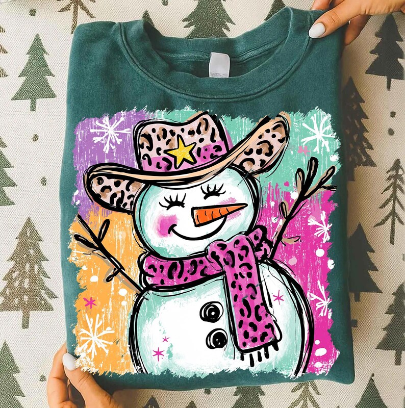 May include: A teal sweatshirt with a playful snowman illustration. The snowman is adorned with a leopard print cowboy hat and scarf, a carrot nose, and twig arms. The background features colourful brushstrokes and snowflakes.