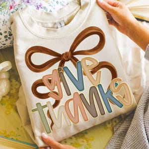 May include: Cream-colored sweatshirt with a brown bow design and the words "GIVE THANKS" in colorful, outlined letters. The sweatshirt is held up by a person.