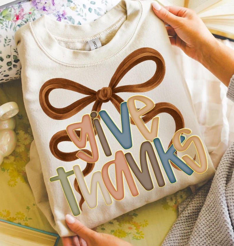 May include: Cream-colored sweatshirt with a brown bow design and the words "GIVE THANKS" in colorful, outlined letters. The sweatshirt is held up by a person.
