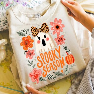 K&ouml;nnte beinhalten: Beiges Sweatshirt mit einem Cartoon-Geist mit Leopardenmuster-Schleife, umgeben von orangefarbenen und rosa Blumen und den Worten "Spooky Season" in Orange. Ein K&uuml;rbis ist ebenfalls im Design enthalten.