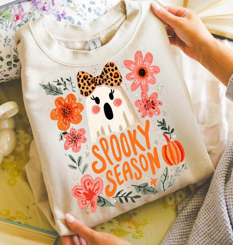 K&ouml;nnte beinhalten: Beiges Sweatshirt mit einem Cartoon-Geist mit Leopardenmuster-Schleife, umgeben von orangefarbenen und rosa Blumen und den Worten "Spooky Season" in Orange. Ein K&uuml;rbis ist ebenfalls im Design enthalten.