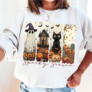 May include: A light gray sweatshirt featuring a Halloween-themed design. The design includes a ghost, haunted house, black cat, pumpkins, and the words "Spooky Season". The artwork is in a vertical rectangular format.