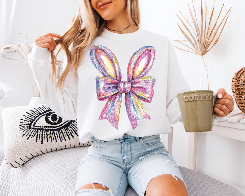 May include: A white sweatshirt with a large, colorful, glittery bow design on the front. The bow is pink, purple, yellow, and blue.
