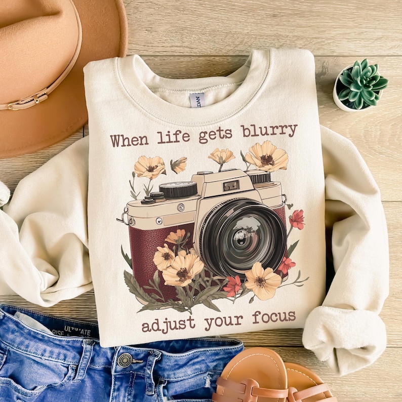 May include: A cream-colored sweatshirt with a vintage camera graphic and the text "When life gets blurry adjust your focus". The camera is surrounded by flowers in shades of red, yellow, and brown.