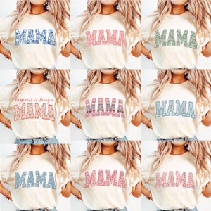 May include: Nine different t-shirts with the word "MAMA" printed on them in various colors and patterns. The shirts are all white and have short sleeves.