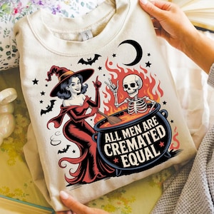 May include: A beige sweatshirt featuring a Halloween-themed graphic. The design includes a witch in a red dress, a skeleton in a cauldron, bats, stars, and the text "ALL MEN ARE CREMATED EQUAL". A crescent moon is also present.