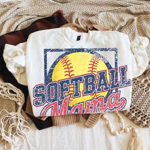 May include: White crew neck sweatshirt with a distressed graphic of a yellow softball, blue and red text that reads "SOFTBALL Mama". The sweatshirt is folded on top of other clothing items.