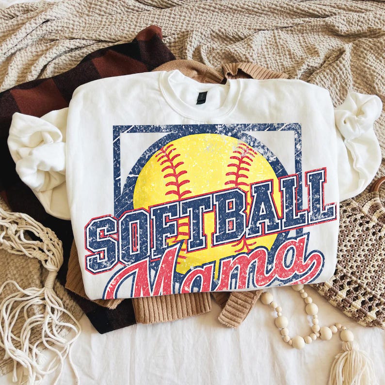 May include: White crew neck sweatshirt with a distressed graphic of a yellow softball, blue and red text that reads "SOFTBALL Mama". The sweatshirt is folded on top of other clothing items.