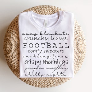 May include: White t-shirt with black text listing fall-themed words: cozy blankets, crunchy leaves, football, comfy sweaters, crackling fires, crispy mornings, pumpkin everything, and chilly nights. The shirt is folded on a woven, circular surface.