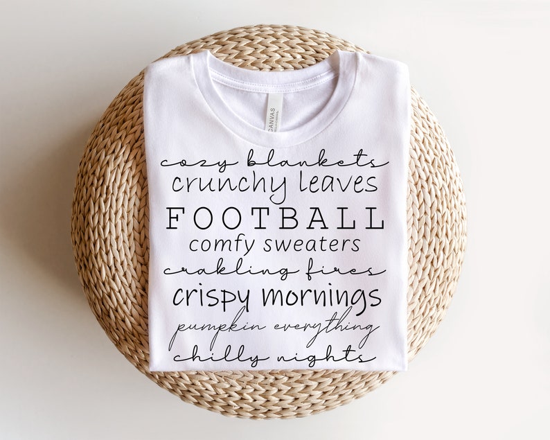 May include: White t-shirt with black text listing fall-themed words: cozy blankets, crunchy leaves, football, comfy sweaters, crackling fires, crispy mornings, pumpkin everything, and chilly nights. The shirt is folded on a woven, circular surface.
