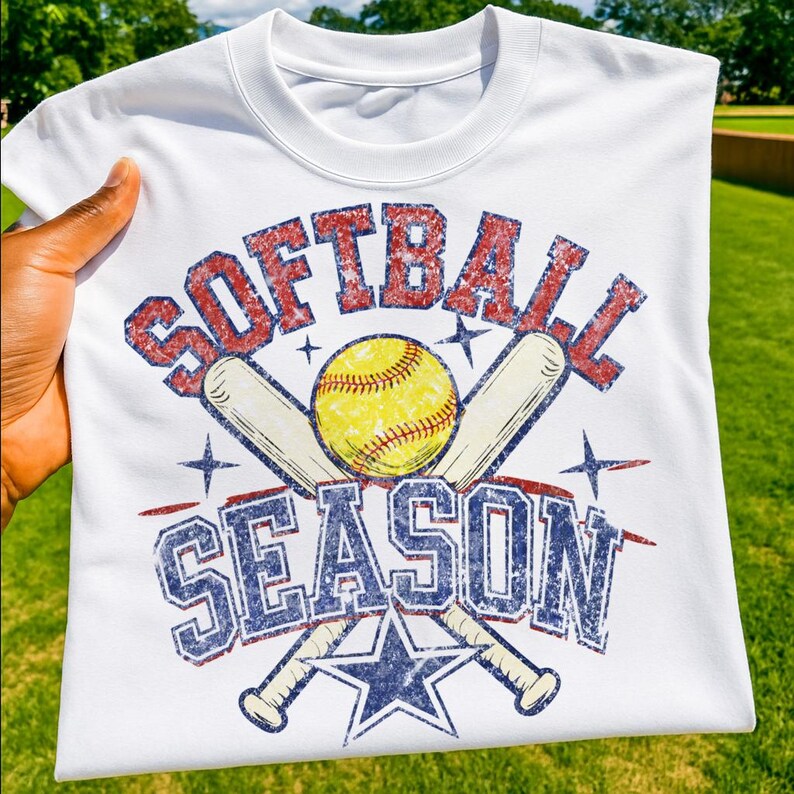 May include: White t-shirt with a distressed graphic design. The design features the words "SOFTBALL SEASON" in red and blue, a yellow softball, crossed baseball bats, and a blue star.