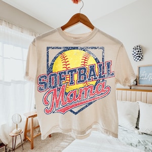 May include: A cream-colored t-shirt with the words "SOFTBALL Mama" in a distressed, vintage style. The design features a yellow softball with red stitching, set within a blue diamond shape. The shirt is hanging on a wooden hanger.