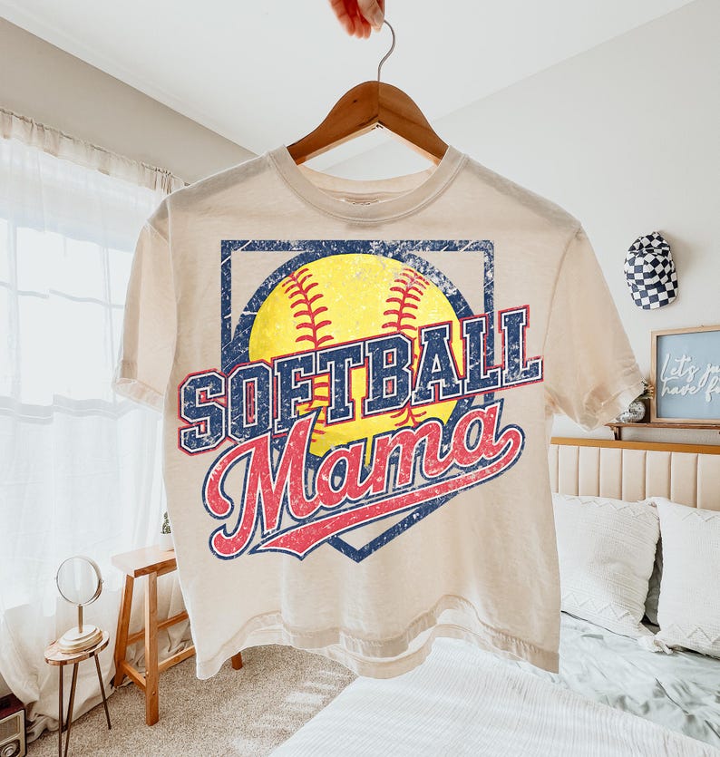 May include: A cream-colored t-shirt with the words "SOFTBALL Mama" in a distressed, vintage style. The design features a yellow softball with red stitching, set within a blue diamond shape. The shirt is hanging on a wooden hanger.