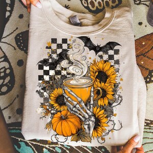 K&ouml;nnte beinhalten: Beiges Sweatshirt mit Halloween-Motiv. Das Design zeigt eine gelbe Kaffeetasse mit Dampf, Sonnenblumen, einen K&uuml;rbis, eine Skeletthand und Flederm&auml;use vor einem karierten Hintergrund. Das Sweatshirt ist gefaltet.