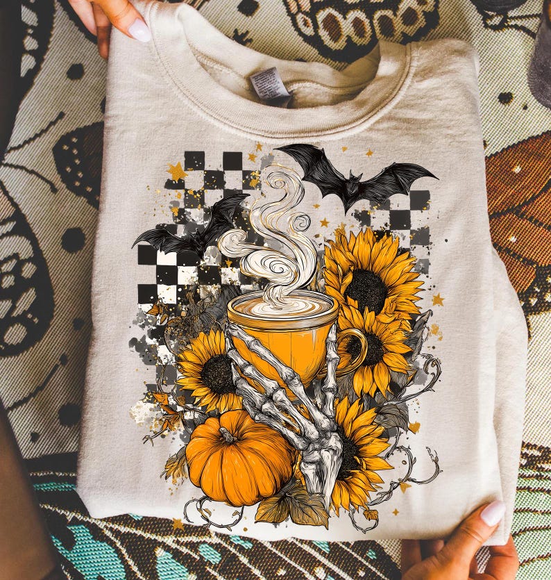 K&ouml;nnte beinhalten: Beiges Sweatshirt mit Halloween-Motiv. Das Design zeigt eine gelbe Kaffeetasse mit Dampf, Sonnenblumen, einen K&uuml;rbis, eine Skeletthand und Flederm&auml;use vor einem karierten Hintergrund. Das Sweatshirt ist gefaltet.
