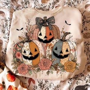 Peut inclure: Sweat-shirt beige avec un motif sur le thème d'Halloween. Le motif comprend trois citrouilles souriantes avec des motifs floraux et à carreaux, entourées de fleurs et de chauves-souris. Les citrouilles ont des yeux et des bouches triangulaires noirs.