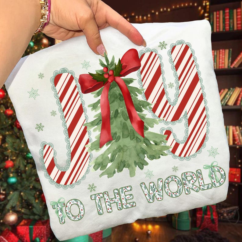 Puede incluir: Coj&iacute;n blanco con tem&aacute;tica navide&ntilde;a con las palabras "JOY TO THE WORLD" en rojo y verde. Las letras "JOY" tienen un dise&ntilde;o de rayas de bast&oacute;n de caramelo con un borde de encaje verde. Un &aacute;rbol de Navidad de acuarela y un lazo rojo est&aacute;n en el centro.