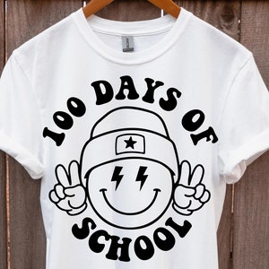 May include: White t-shirt with a black graphic design of a smiley face wearing a hat and peace signs. The text "100 Days of School" is written in a retro font around the smiley face.