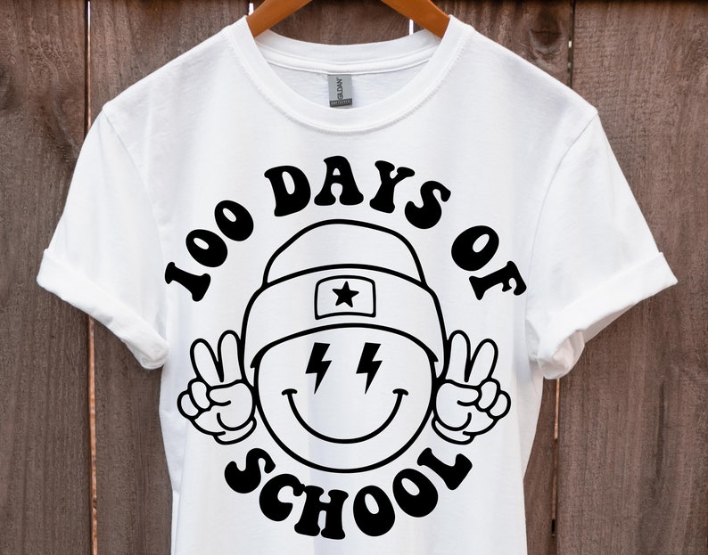 May include: White t-shirt with a black graphic design of a smiley face wearing a hat and peace signs. The text "100 Days of School" is written in a retro font around the smiley face.