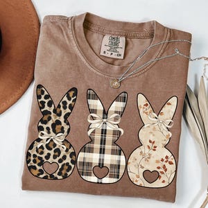 May include: A brown t-shirt with three bunny silhouettes. The bunnies are decorated with leopard print, plaid, and floral patterns. Each bunny has a heart cutout and a bow.