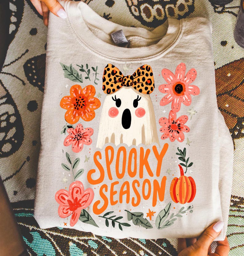K&ouml;nnte beinhalten: Hellbeige Sweatshirt mit einem Geist-Design mit Leopardenmuster-Schleife, umgeben von orangefarbenen und rosa Blumen, gr&uuml;nen Bl&auml;ttern und einem K&uuml;rbis. Die Worte "Spooky Season" sind in Orange geschrieben.