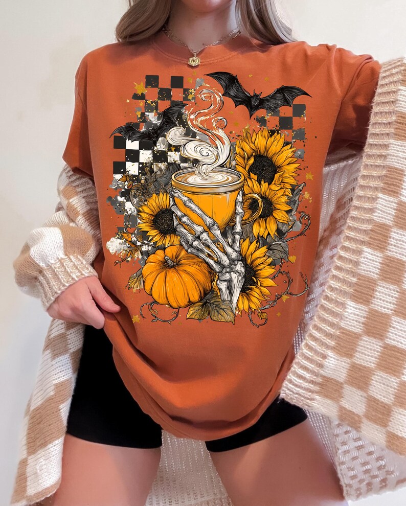 K&ouml;nnte beinhalten: Orangefarbenes T-Shirt mit einem Halloween-Motiv. Die Grafik zeigt eine Kaffeetasse, die von einer Skeletthand gehalten wird, Sonnenblumen, einen K&uuml;rbis und Flederm&auml;use. Das T-Shirt hat einen Rundhalsausschnitt und kurze &Auml;rmel.