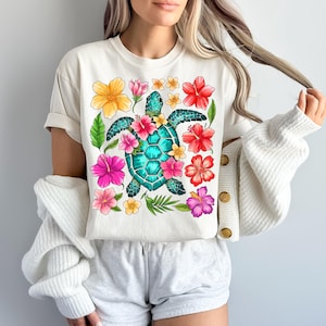 May include: White t-shirt with a colourful floral design featuring a green sea turtle with a blue shell. The flowers are pink, red, yellow, and orange.