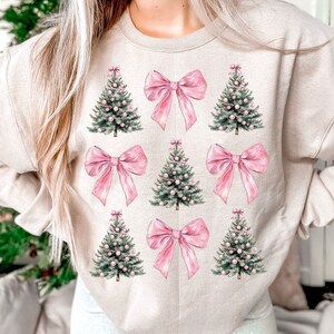 May include: Beige sweatshirt with a repeating pattern of pink bows and Christmas trees. The trees are green with pink baubles, and the bows are a solid pink colour. The design is centred on the front of the sweatshirt.