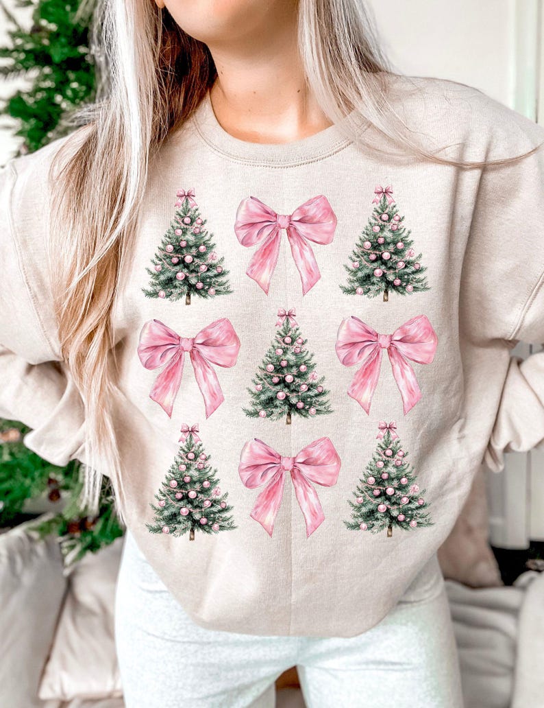 May include: Beige sweatshirt with a repeating pattern of pink bows and Christmas trees. The trees are green with pink baubles, and the bows are a solid pink colour. The design is centred on the front of the sweatshirt.