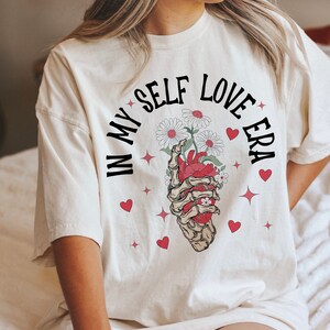 May include: Cream-colored t-shirt with the text "IN MY SELF LOVE ERA" in black, arched above a graphic. The graphic features a skeletal hand holding a red heart and white daisies. Red hearts and starbursts are scattered around the design.