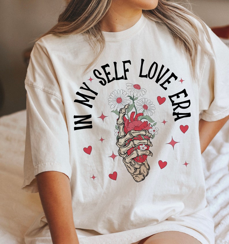 May include: Cream-colored t-shirt with the text "IN MY SELF LOVE ERA" in black, arched above a graphic. The graphic features a skeletal hand holding a red heart and white daisies. Red hearts and starbursts are scattered around the design.