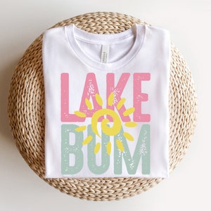 Op de afbeelding: Wit T-shirt met een roze, gele en groene afbeelding met de tekst "Lake Bum".