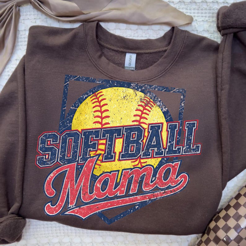 May include: A brown sweatshirt featuring a yellow softball with red stitching, set within a blue diamond-shaped frame. The words "SOFTBALL Mama" are printed in a distressed, vintage style, with blue and red lettering.