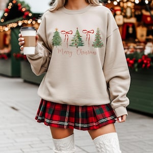 May include: Beige sweatshirt with a watercolor design of Christmas trees and red bows, with the words "Merry Christmas" in red script. The person is wearing a red and green plaid skirt.