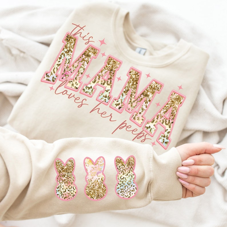 以下が含まれることがあります： ベージュのトレーナーに、ピンクとゴールドのグリッター文字で「This Mama loves her peeps」と書かれています。袖には、ピンクとゴールドのグリッターでできたピープの模様が3つあります。