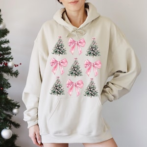 May include: A beige hoodie with a Christmas-themed design. The design features green Christmas trees and pink bows. The trees have white accents, and the bows are a soft pink. The hoodie has a drawstring hood and a front pocket. The design is centered on the front.