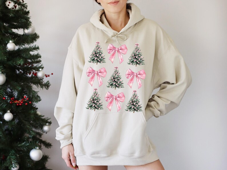 May include: A beige hoodie with a Christmas-themed design. The design features green Christmas trees and pink bows. The trees have white accents, and the bows are a soft pink. The hoodie has a drawstring hood and a front pocket. The design is centered on the front.