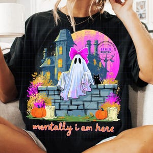 Puede incluir: Una camiseta negra con un estampado gr&aacute;fico de un fantasma blanco con un lazo rosa brillante, sentado sobre un muro de piedra frente a una casa oscura, un gato negro, calabazas naranjas, velas y el texto "mentally i am here" en letras naranjas.
