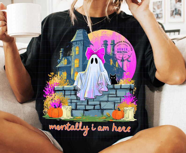 Puede incluir: Una camiseta negra con un estampado gr&aacute;fico de un fantasma blanco con un lazo rosa brillante, sentado sobre un muro de piedra frente a una casa oscura, un gato negro, calabazas naranjas, velas y el texto "mentally i am here" en letras naranjas.